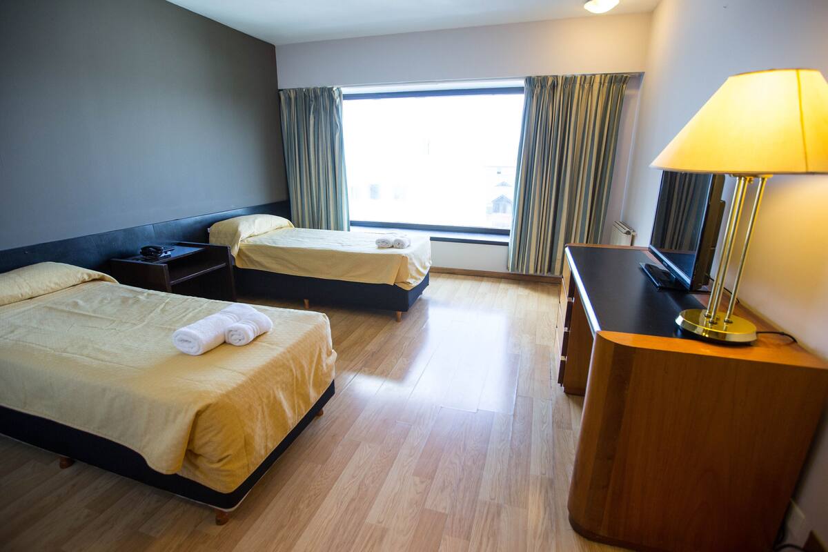 Une chambre d’hôtel avec deux lits, un bureau, une lampe et une grande fenêtre avec des rideaux.