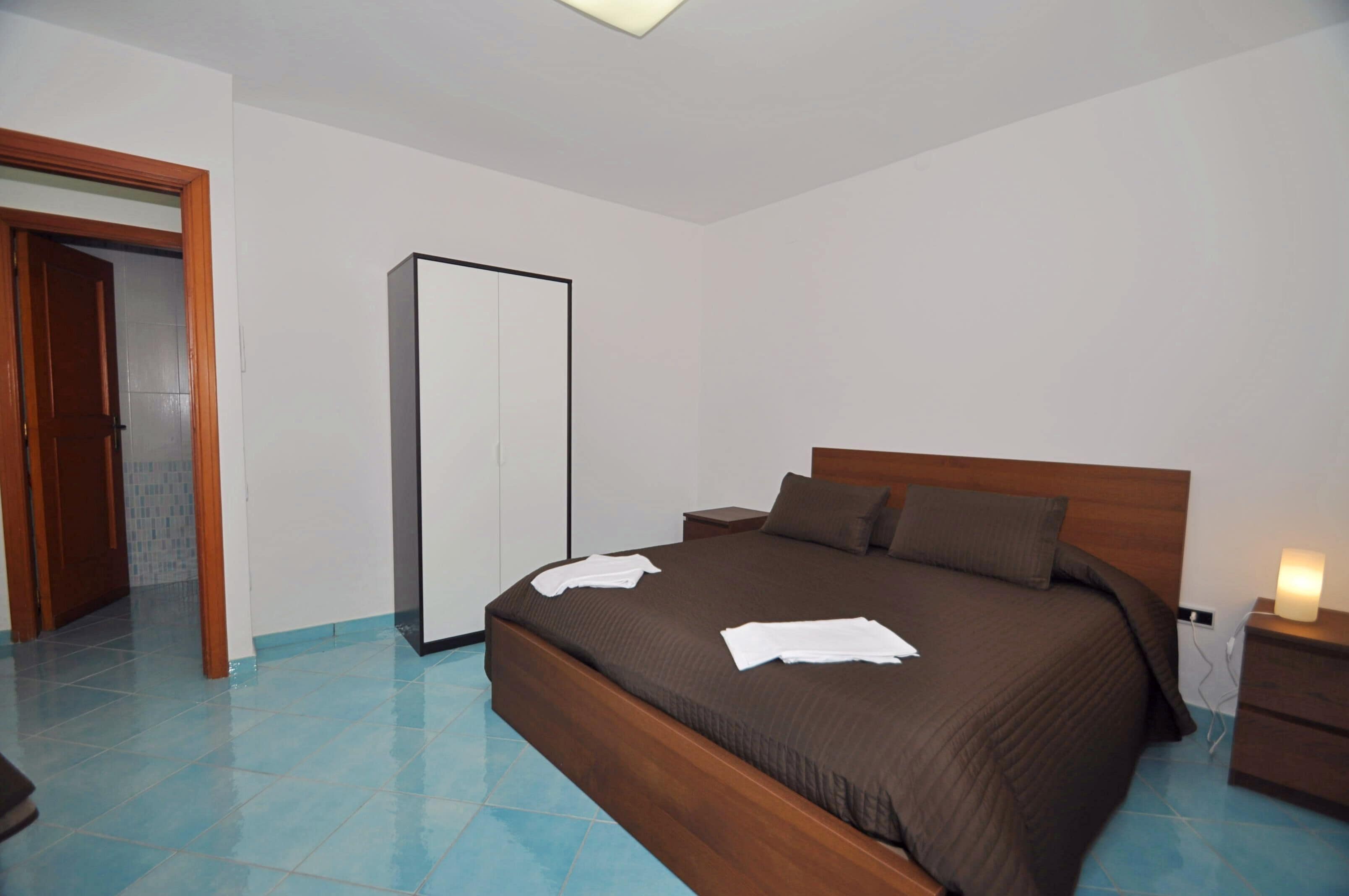 Un dormitorio con cama de madera, mesita de noche, lámpara de noche, armario y suelo de baldosas.