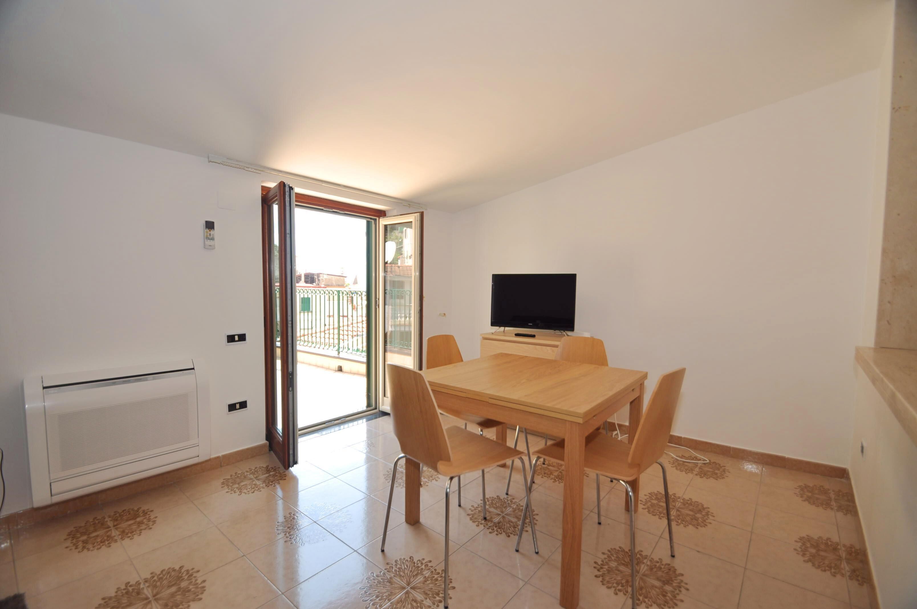 Un espace repas comprenant une table et des chaises, un téléviseur et une porte-fenêtre coulissante donnant sur un balcon extérieur.