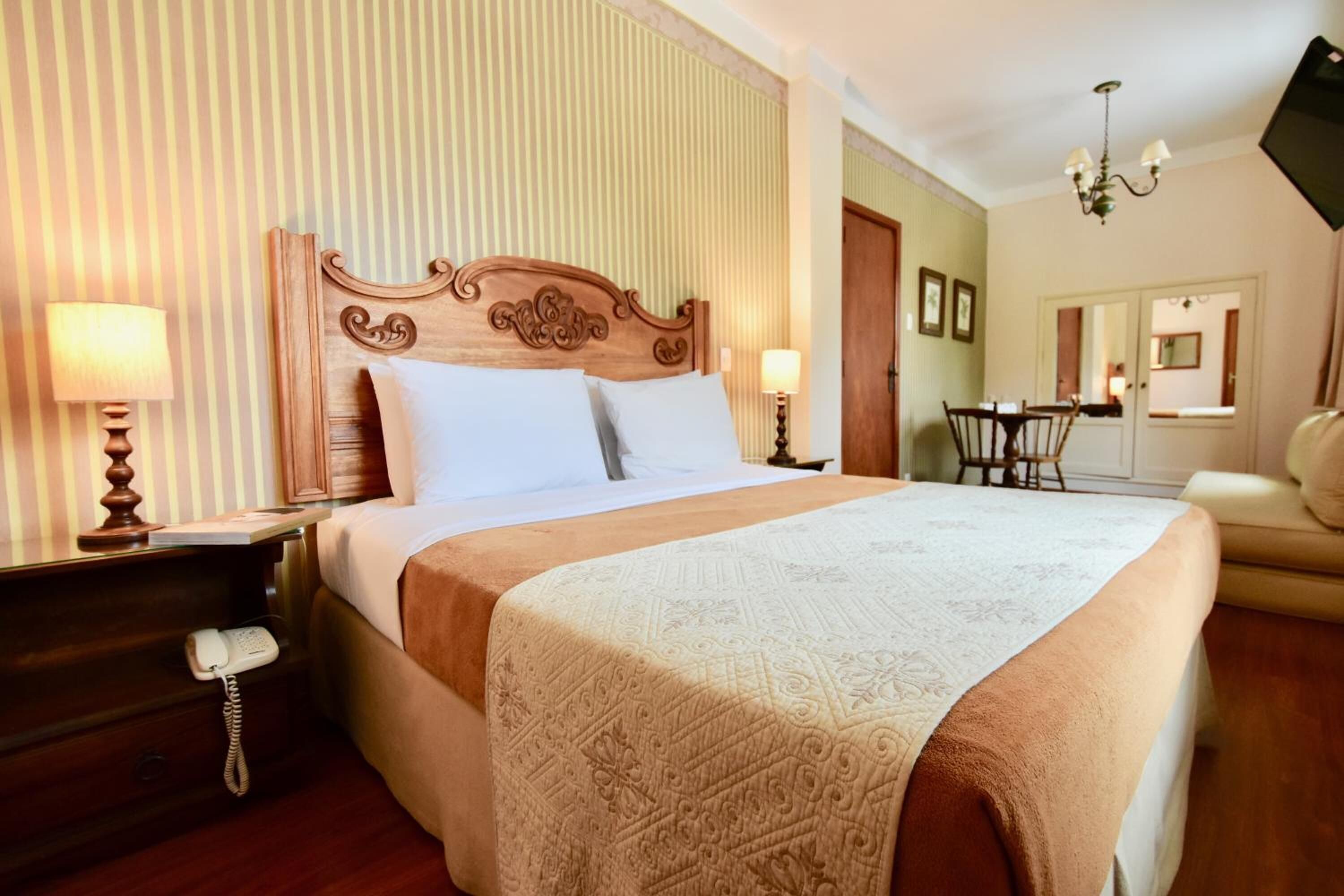A hotel room with a large bed, wooden headboard, two bedside lamps, a telephone, and a dining area with a table and chairs.
