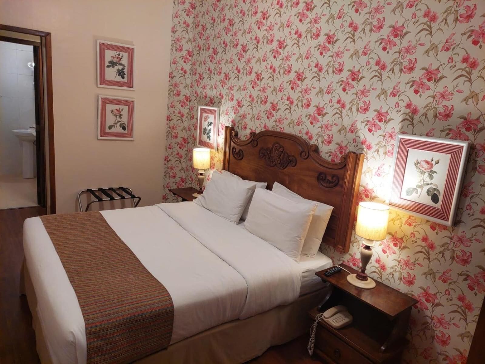 A neatly made bed with white linens and a brown striped blanket, a wooden headboard, two bedside lamps, a nightstand with a telephone, and floral wallpaper.