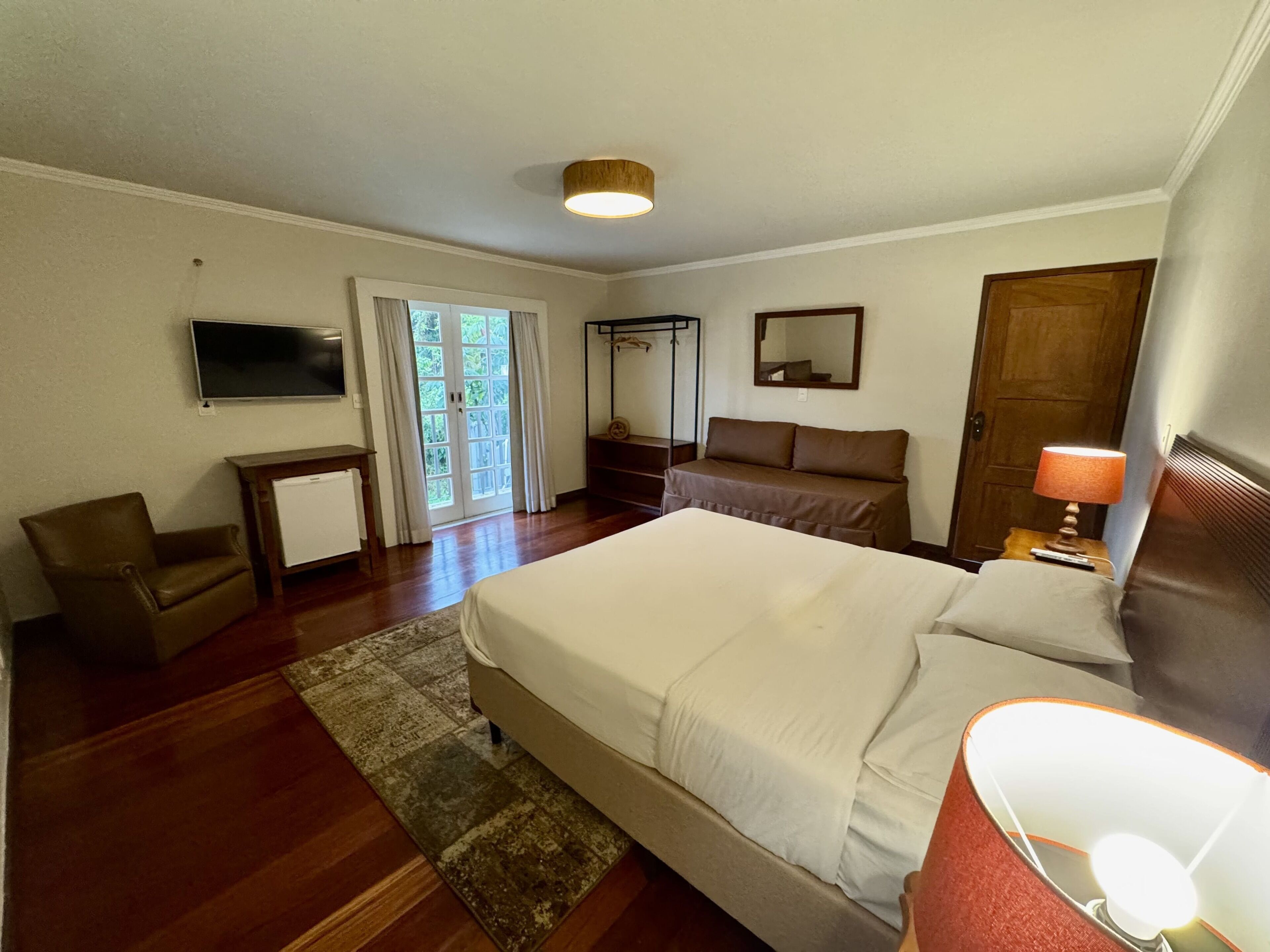 A bedroom with a bed, a chair, a desk, a TV, a mirror, and a door.