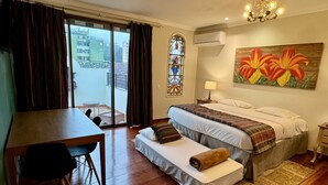 Une chambre d’hôtel comprenant un lit, un bureau, une chaise, une fenêtre avec des rideaux, un vitrail, un appareil de climatisation et un tableau représentant des fleurs.