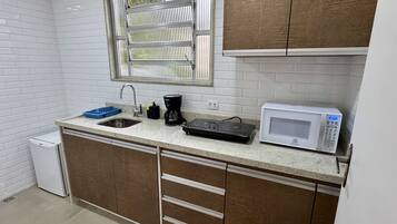 Een compacte keuken met witte betegelde wanden, een raam met zonwering, een spoelbak, een koffiezetapparaat, een magnetron en een kleine koelkast.