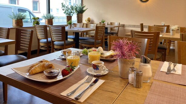 A dining area with wooden tables and chairs, breakfast items like croissants, strawberries, and orange juice, and potted plants. - Hotel Russmann (Goldbach)