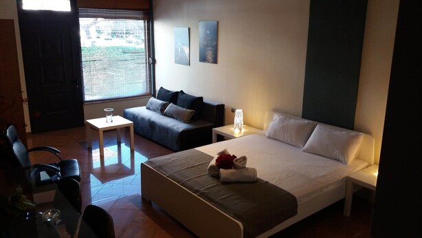 Habitación de hotel con cama, sofá, una mesita y una silla. Hay dos cuadros en la pared y una ventana con persianas.