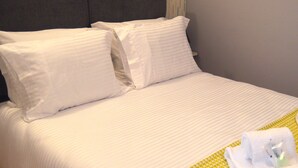 Una cama bien hecha con ropa blanca y almohadas, una manta de color verde con estampado y una toalla blanca doblada sobre la cama.