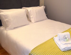 Una cama bien hecha con ropa blanca y almohadas, una manta de color verde con estampado y una toalla blanca doblada sobre la cama.