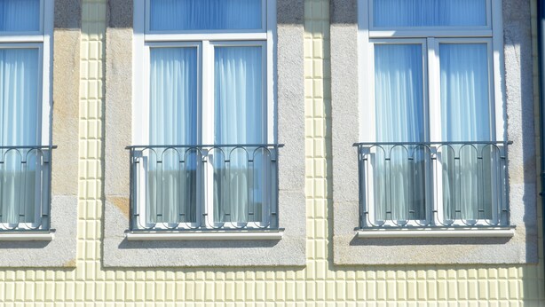 Fachada de edificio con tres ventanas, cada una con balcón y cortinas blancas.