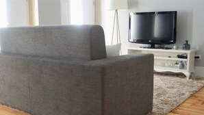 Un sofá gris en una sala de estar con un televisor de pantalla plana sobre un soporte blanco, una lámpara y una mesita con objetos decorativos.