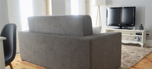 Un sofá gris en una sala de estar con un televisor de pantalla plana sobre un soporte blanco, una lámpara y una mesita con objetos decorativos.