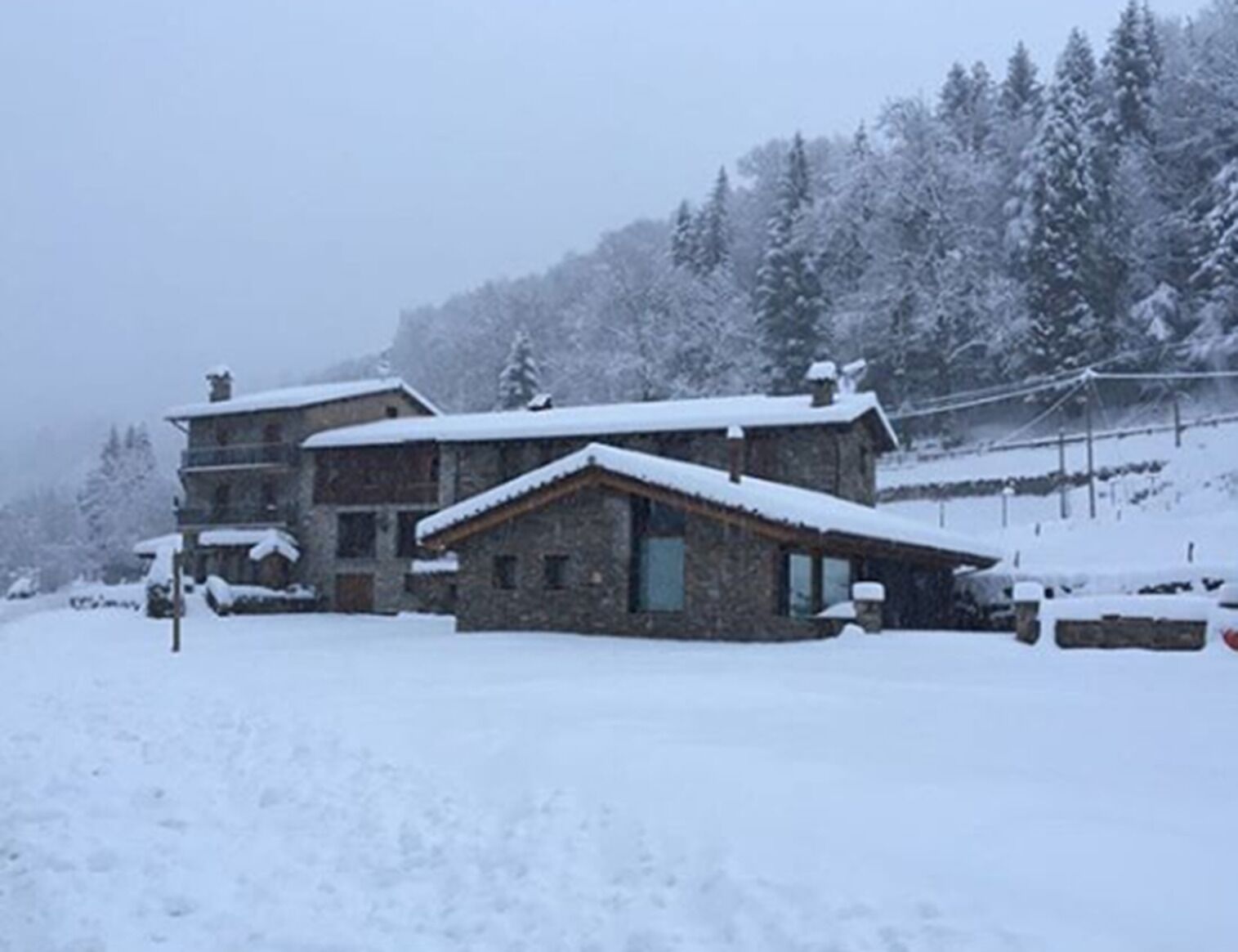 A stone building with a snow-covered roof and multiple windows, surrounded by snow-covered trees and a hilly landscape.
