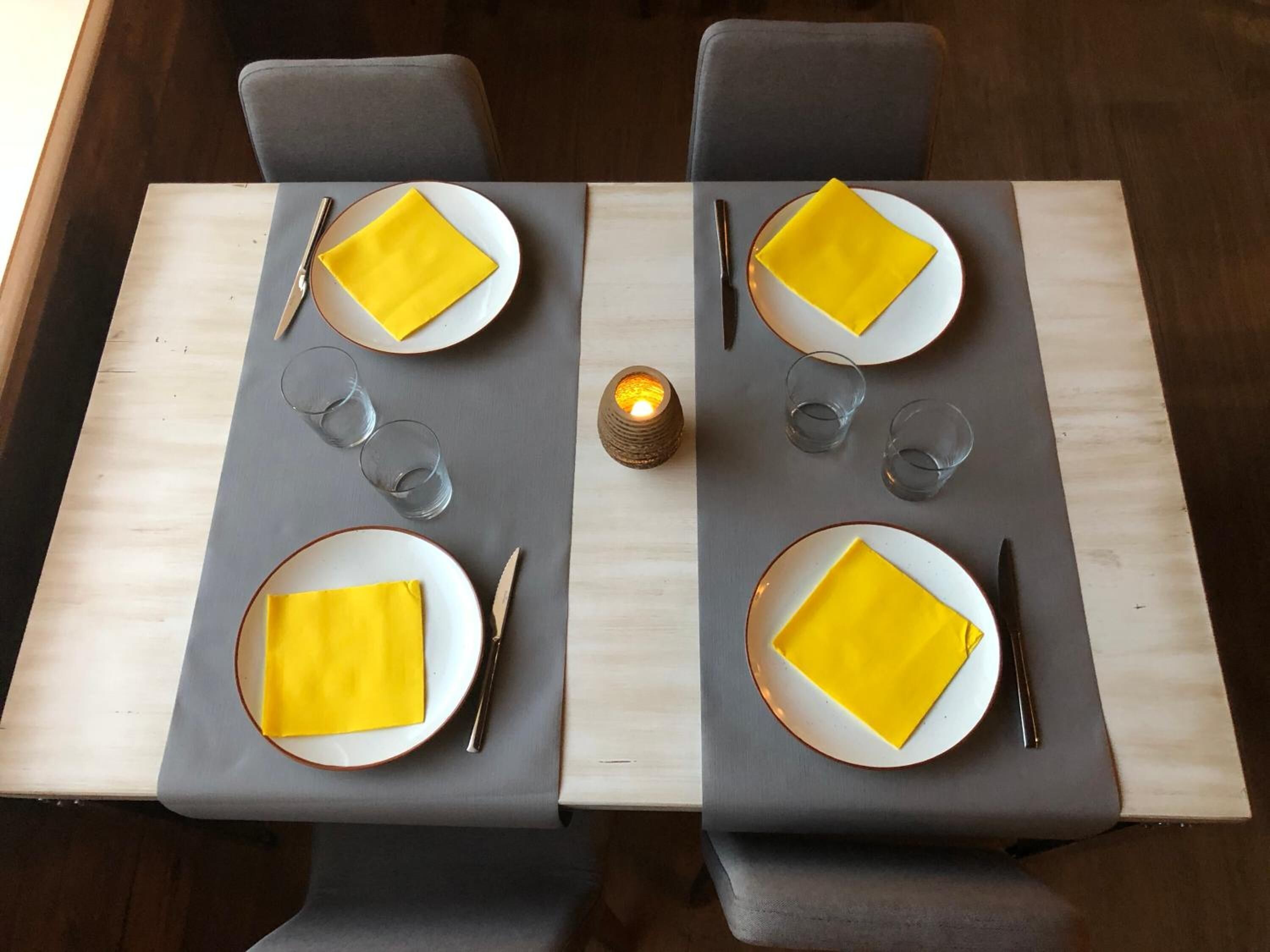 A dining table set with yellow napkins, plates, cutlery, and glasses, centered by a candle.