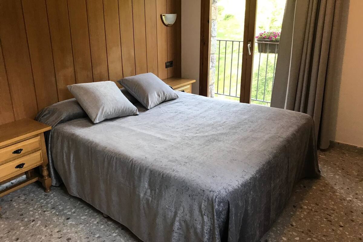 A bedroom with a large bed, wooden panel walls, a nightstand, and a view of a balcony with plants.