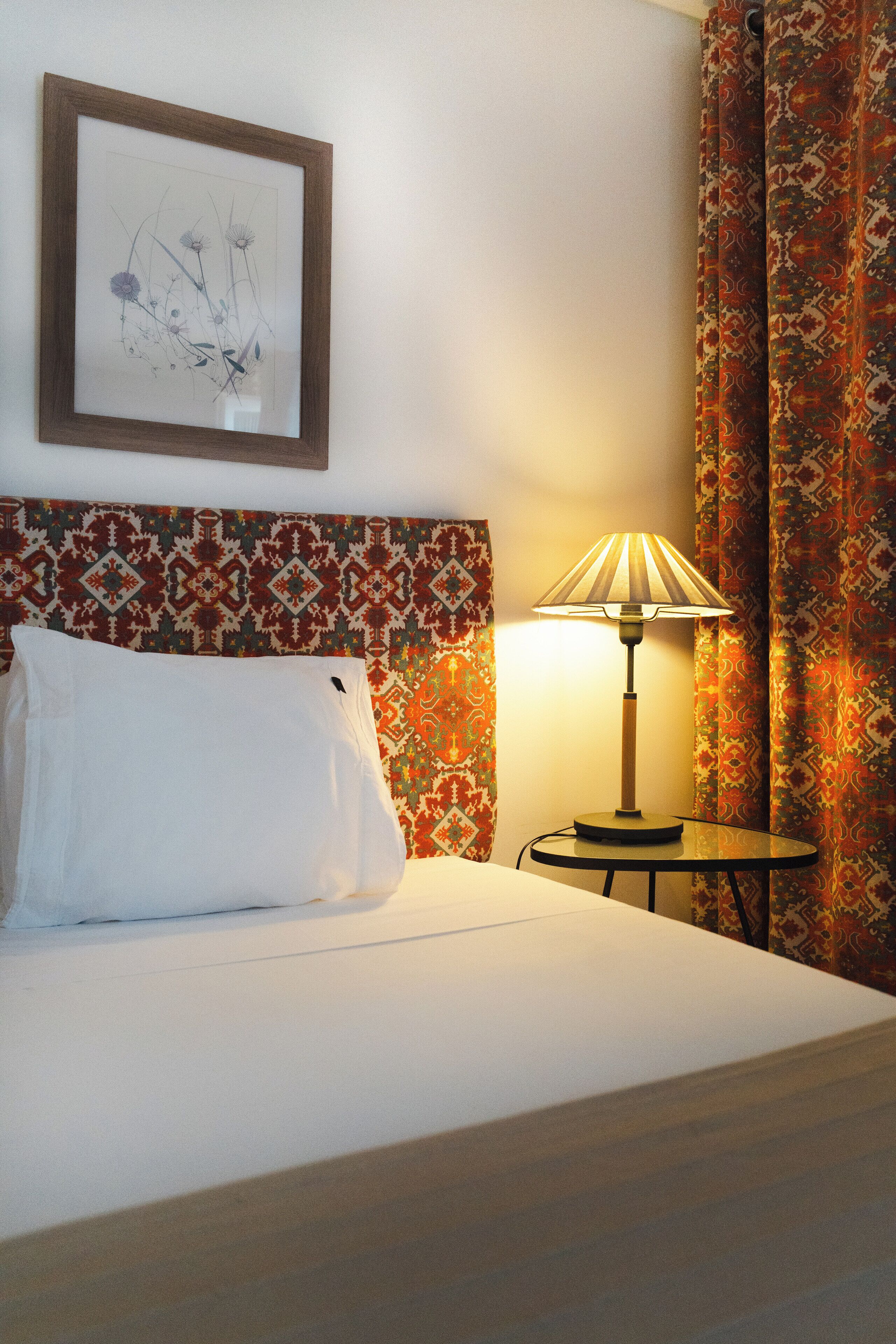 A hotel room with a bed, a bedside lamp, a framed artwork on the wall, and patterned curtains.