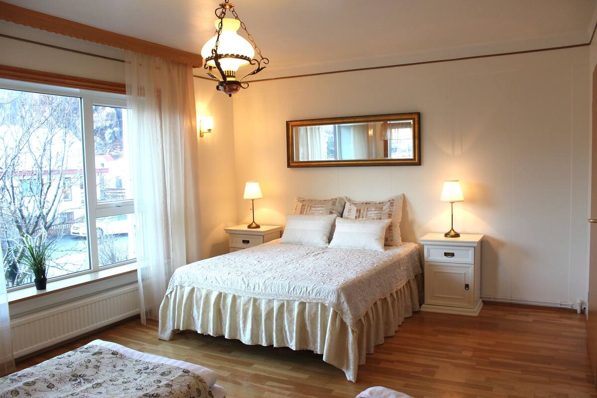 Une chambre à coucher avec un grand lit, deux tables de chevet avec des lampes, une fenêtre avec des rideaux et un miroir au mur.