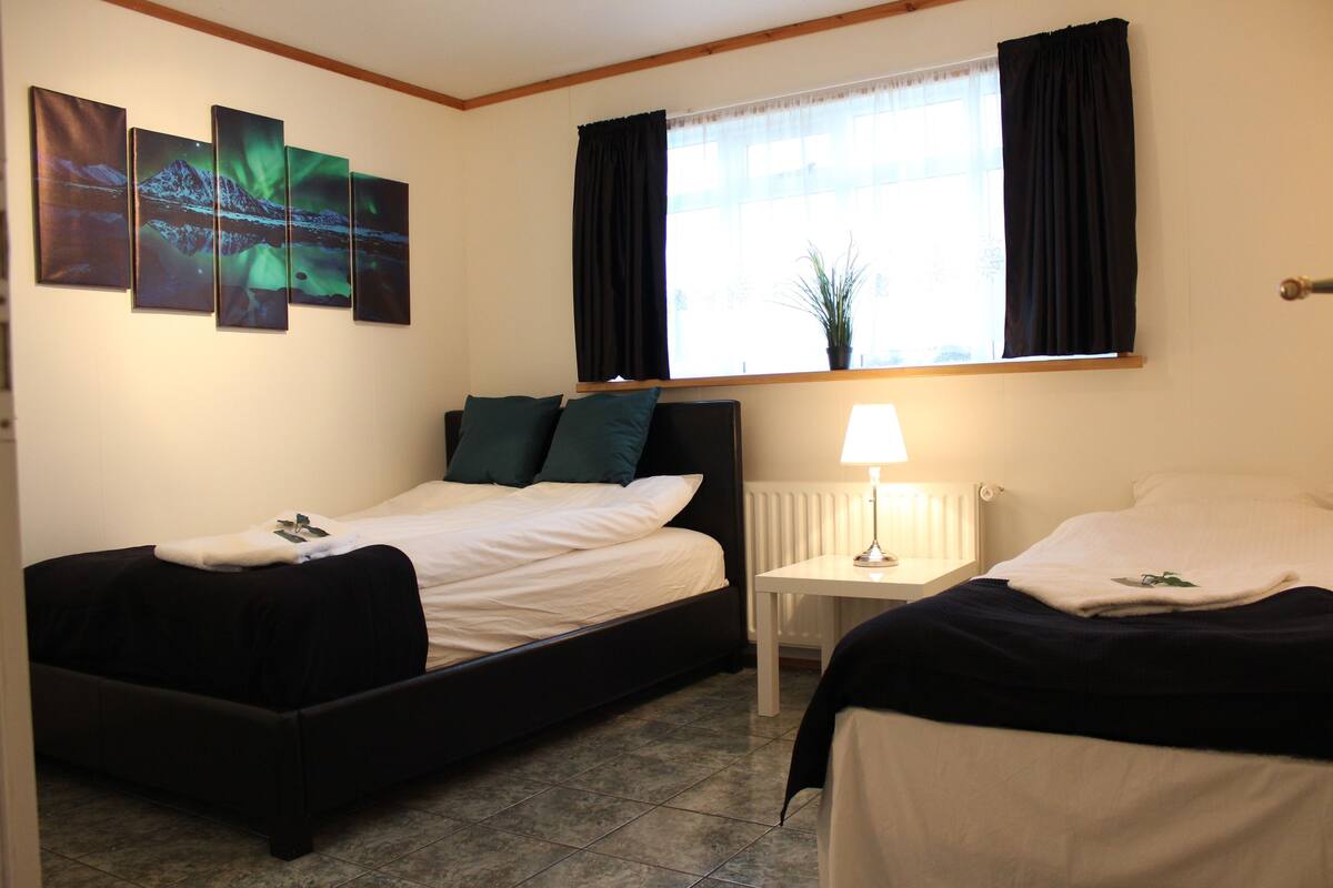 Une chambre d’hôtel avec deux lits, une table de chevet, une fenêtre avec des rideaux et un mur orné de quatre tableaux.