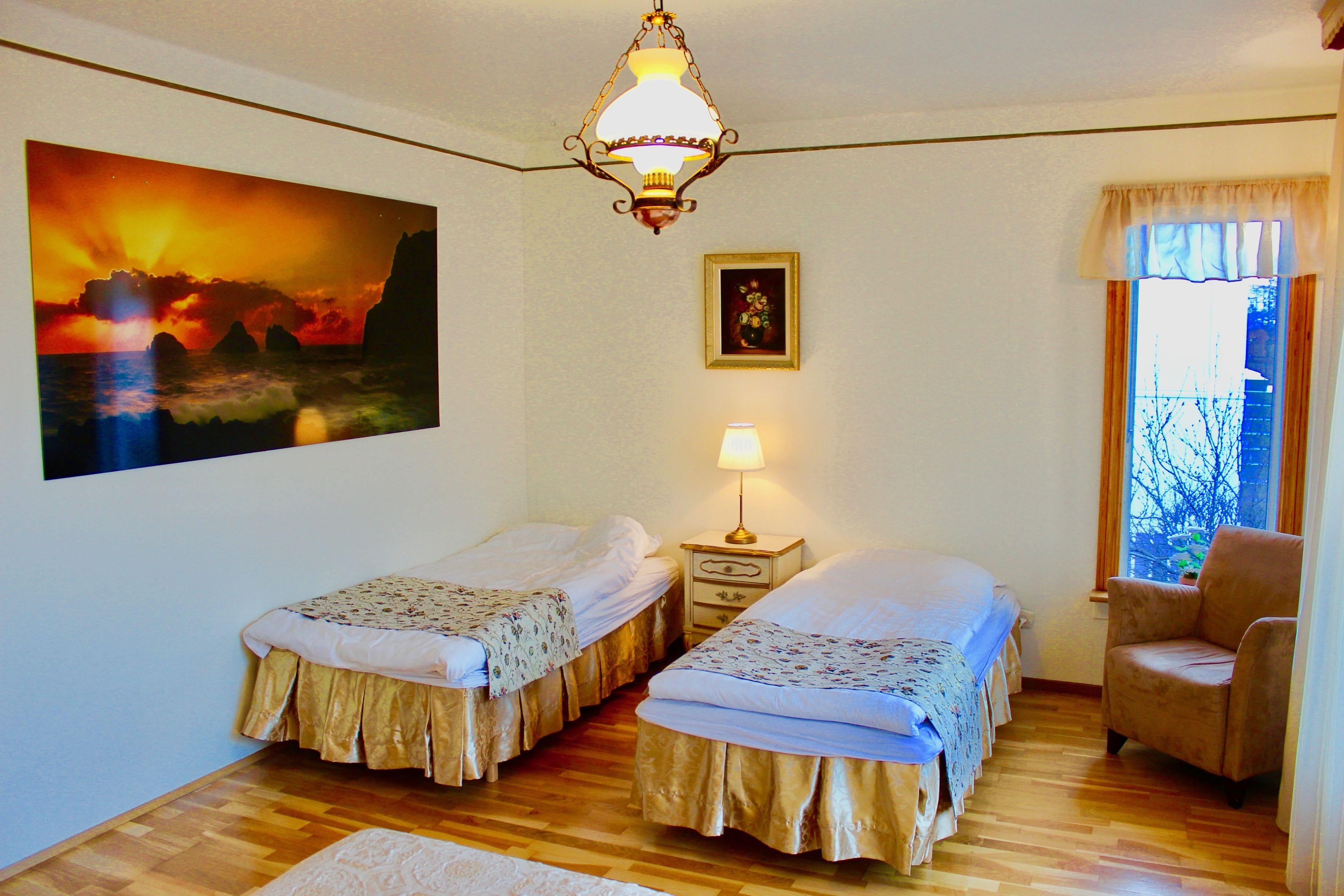 A room with two single beds, a nightstand with a lamp, a chair, a painting of a sunset, and a window with curtains.