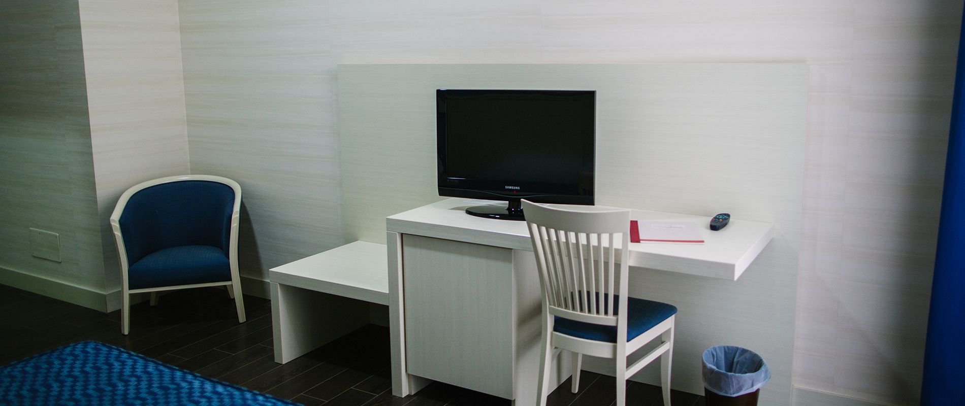 A small, clean room with a white desk, a television, a blue chair, and a trash bin.