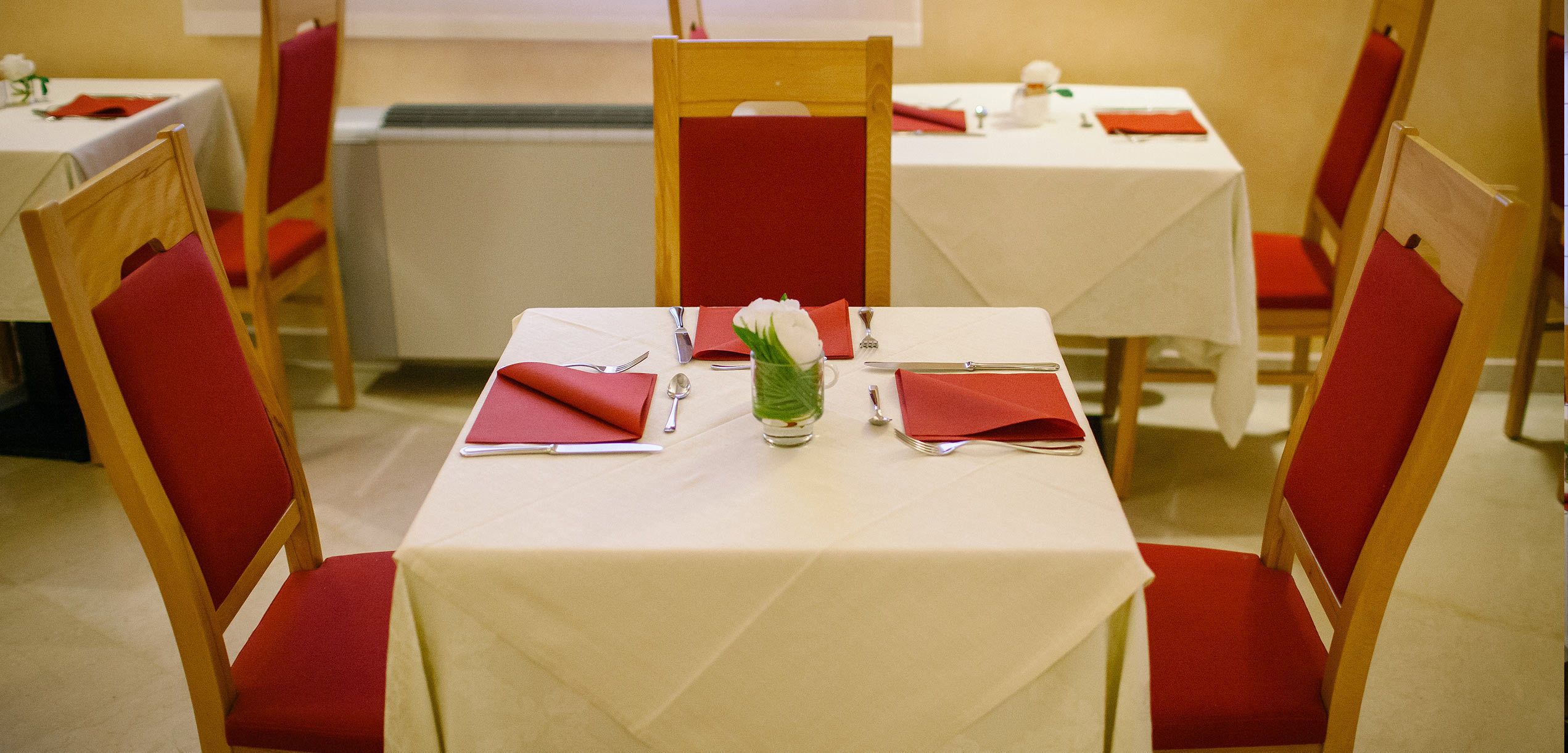 A dining area with tables set for two, each with red chairs, white tablecloths, and red napkins.