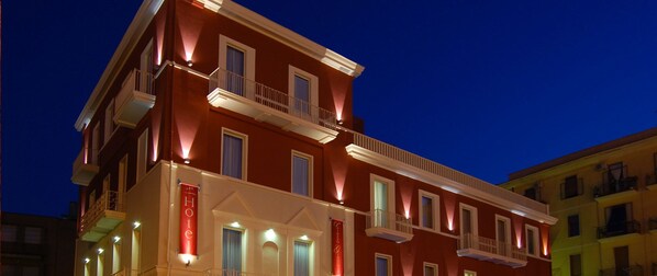 A multi-story hotel with balconies, a red facade, and illuminated signage indicating it is a hotel. - Hotel Palazzo Giancola (San Severo)