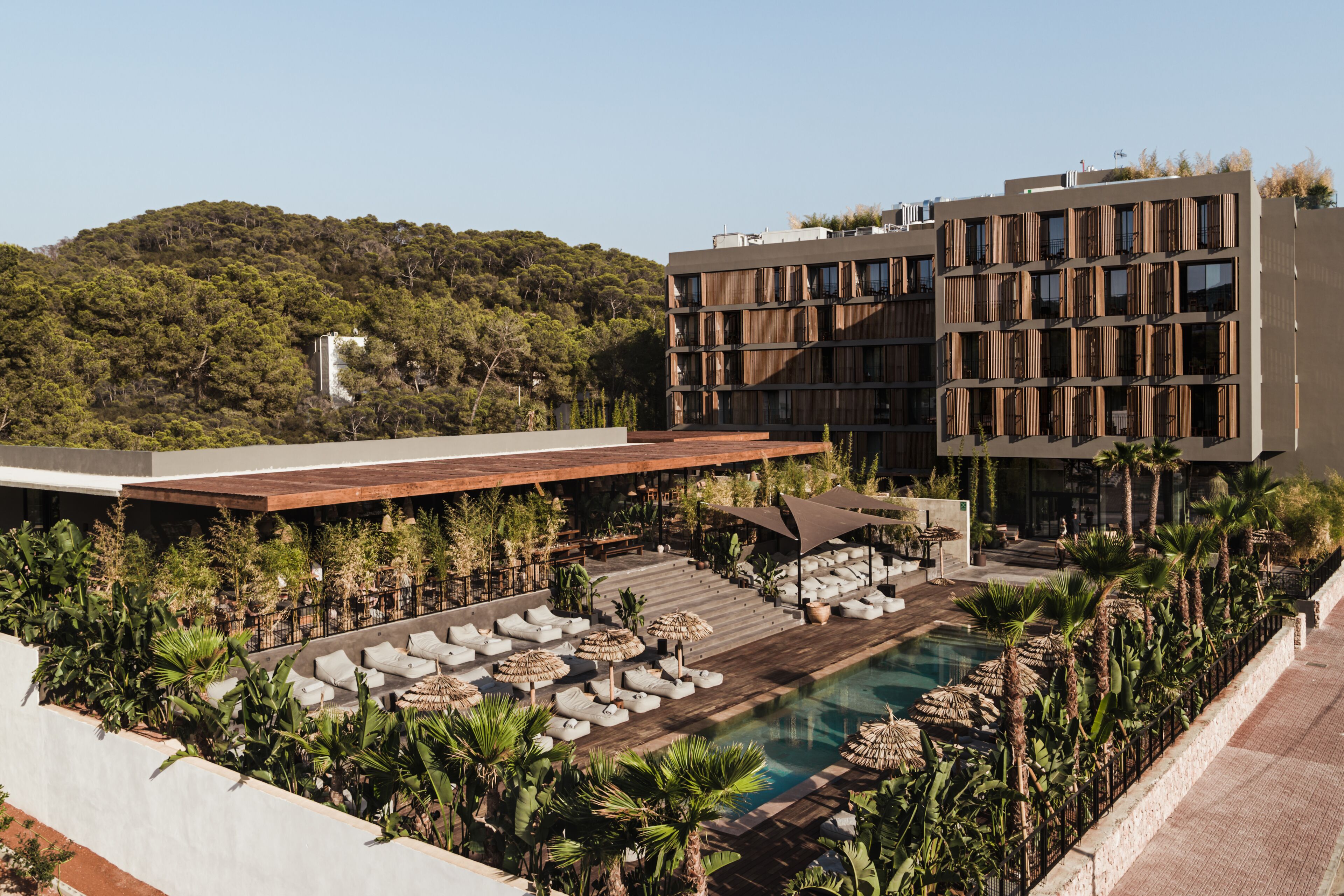A modern hotel with a rooftop pool area, surrounded by lush greenery and a view of a forested hill.