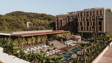 A modern hotel with a rooftop pool area, surrounded by lush greenery and a view of a forested hill.