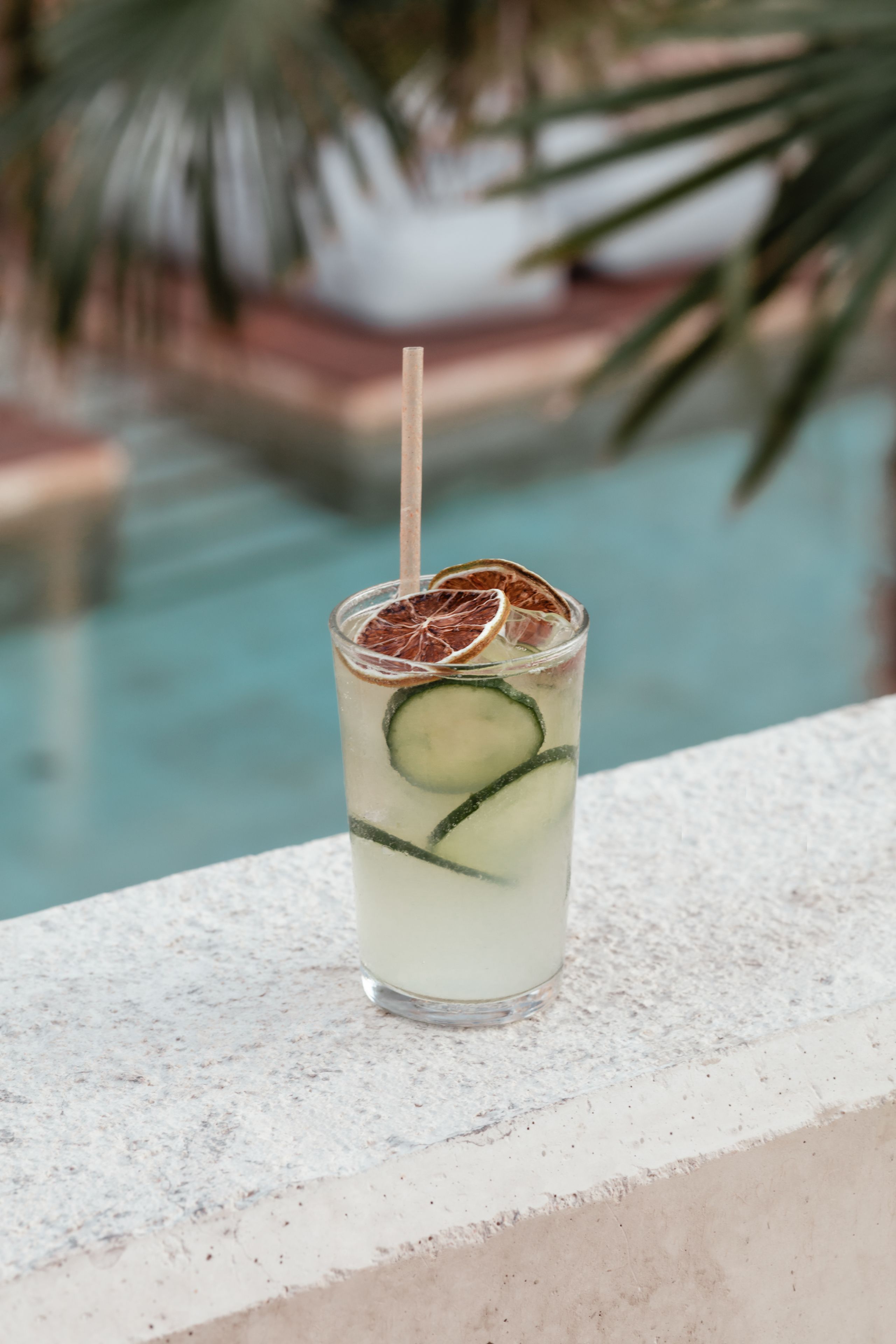 Un verre transparent avec une paille, contenant une boisson agrémentée de rondelles de concombre et de fruits secs, posé sur une surface texturée près d’une piscine.