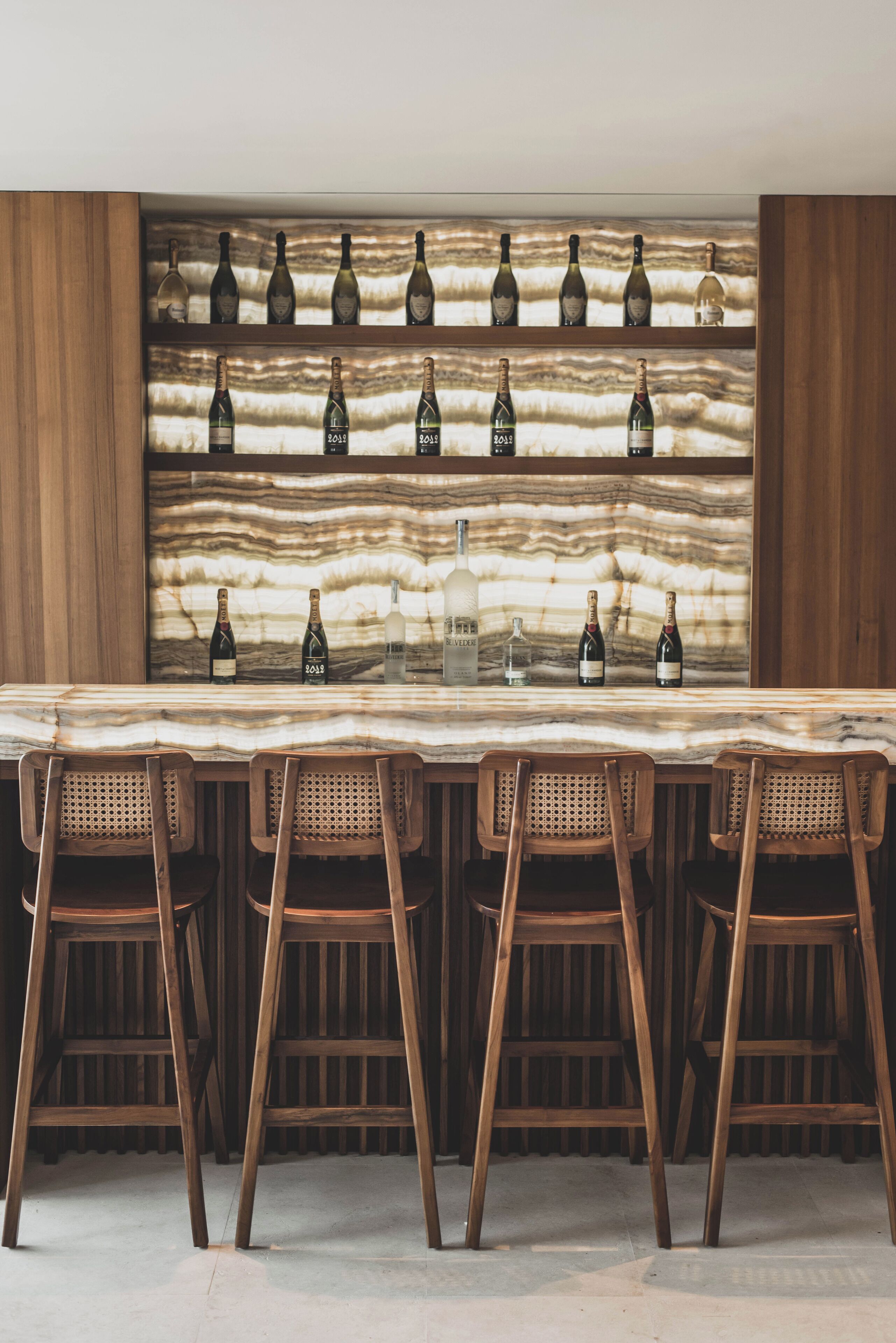 Una zona de bar con mostrador de mármol, sillas de madera y una estantería con botellas de vino.