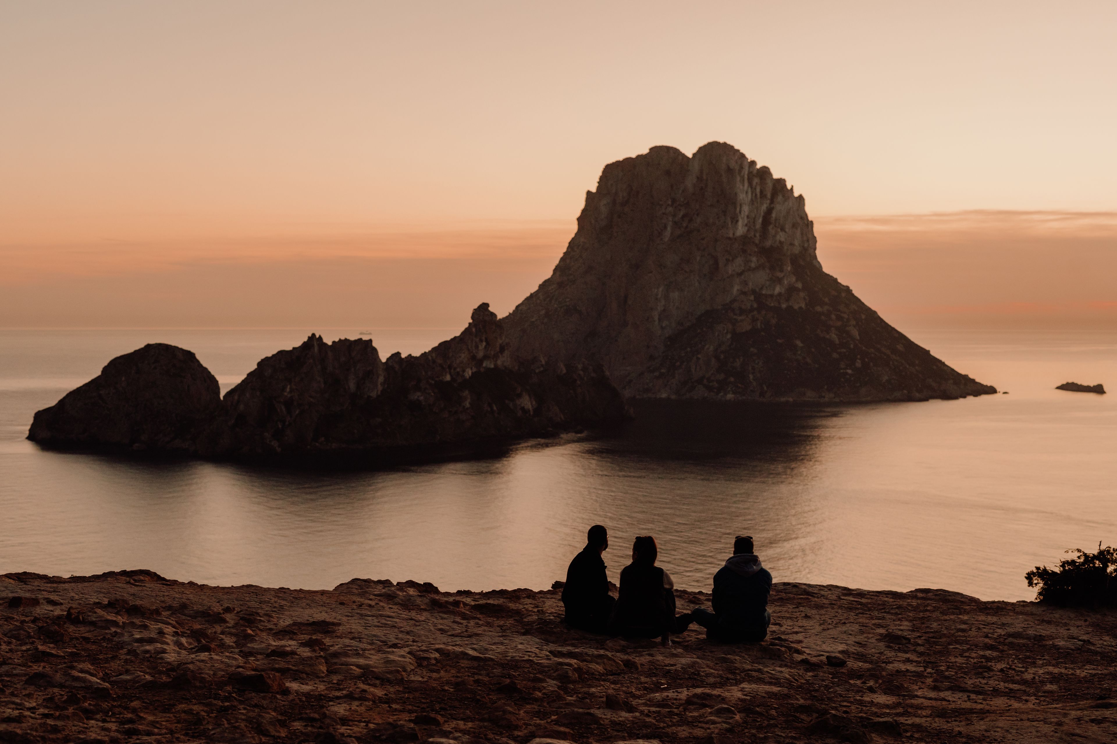 A rocky coastline with three silhouetted figures sitting on the shore, overlooking a calm sea and a prominent rock formation.