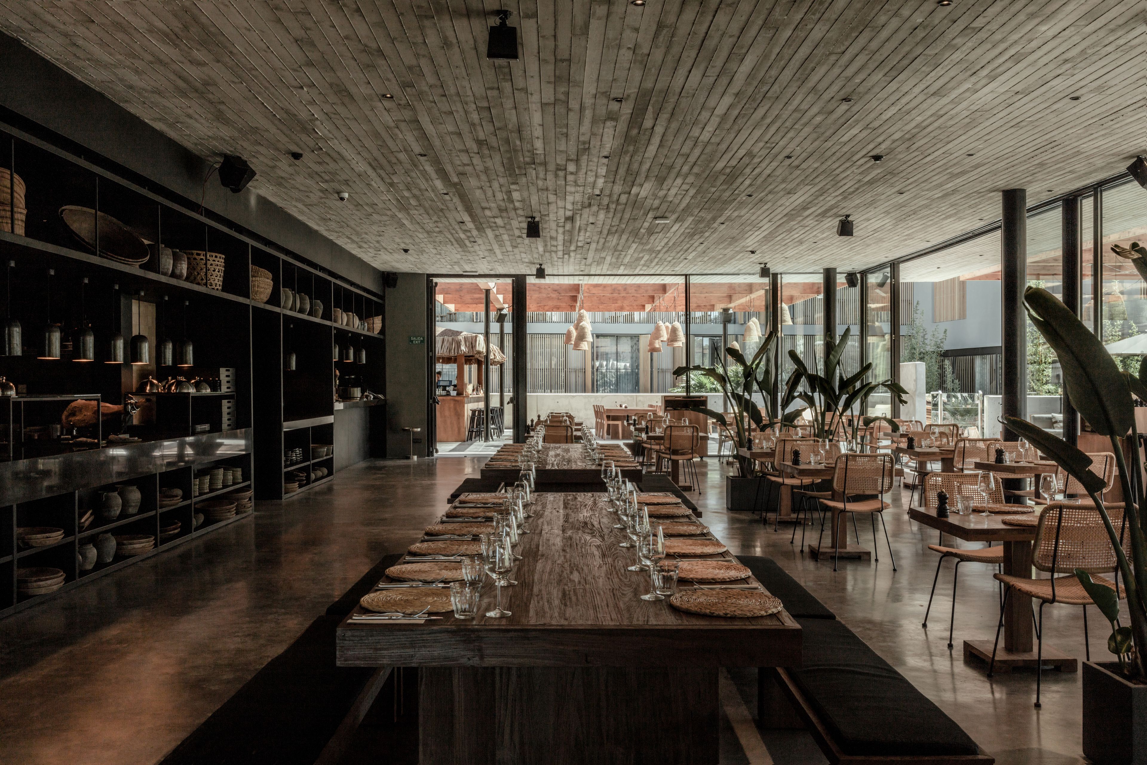 Un comedor amplio con una larga mesa de madera dispuesta para una comida, rodeado de sillas y estantes con utensilios de cocina.