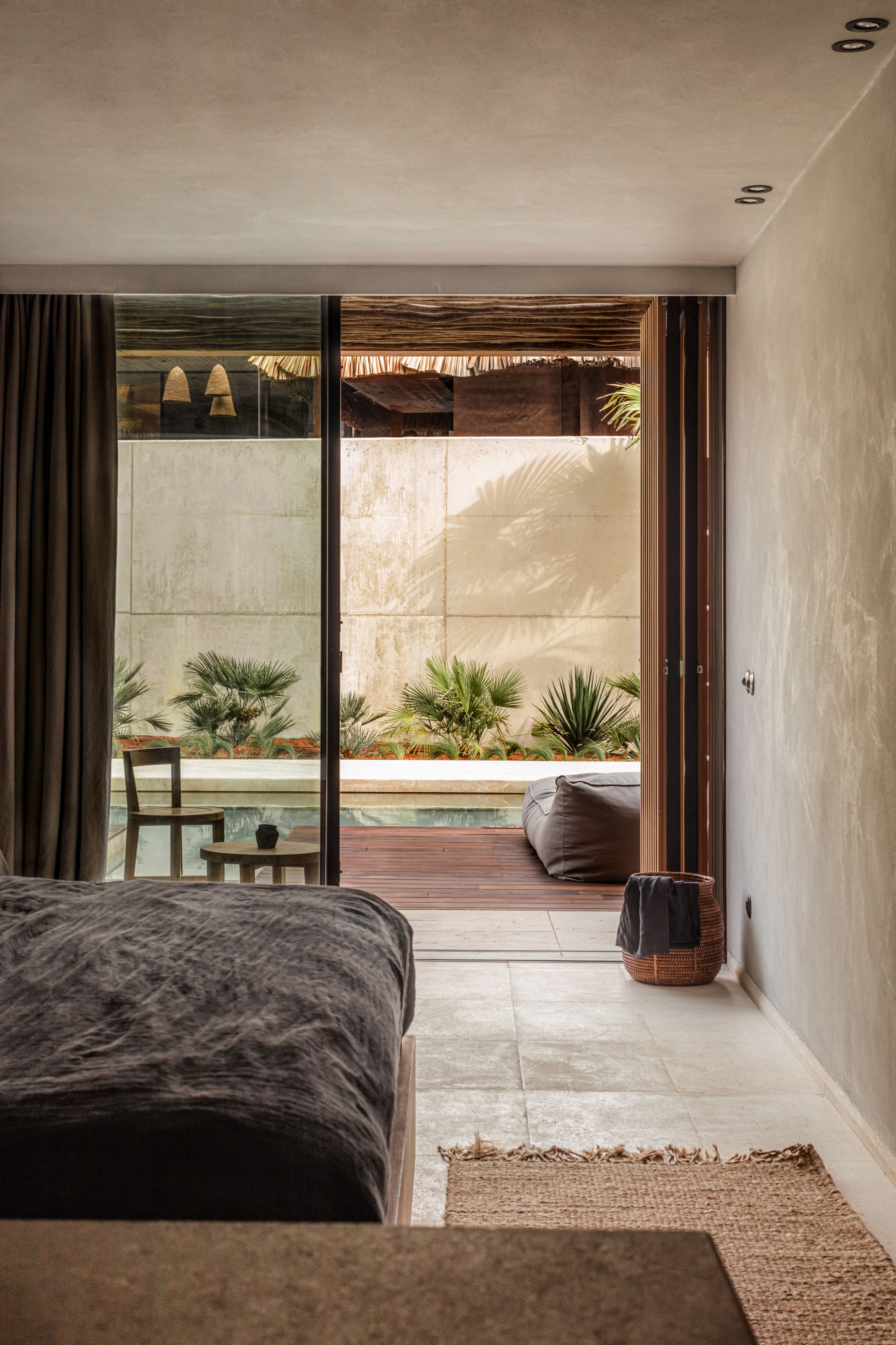 Un dormitorio con una cama grande, una mesita y una silla. Hay una puerta corrediza de vidrio que da a un balcón con plantas y vistas a un edificio.