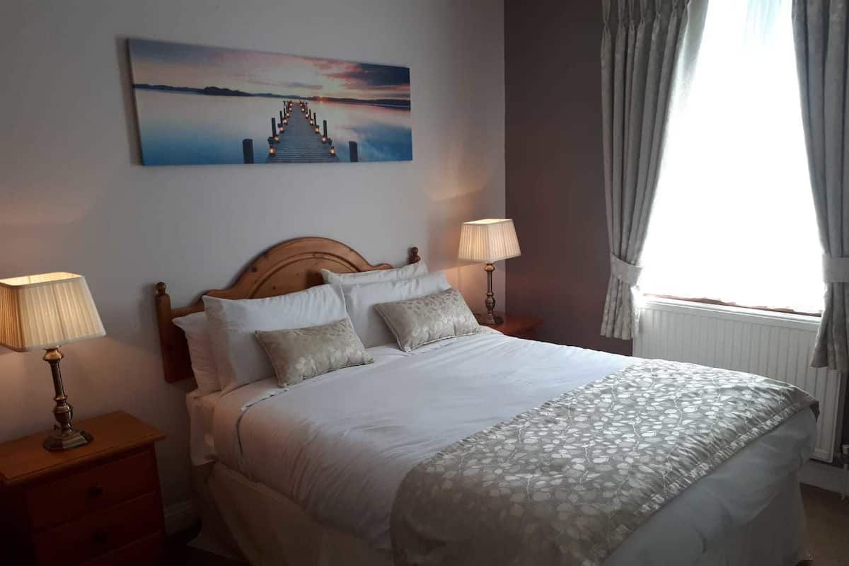 Ein ordentlich gemachtes Bett mit einem hölzernen Kopfteil, zwei Nachttischlampen und einem Gemälde eines Sonnenuntergangs über dem Wasser an der Wand.