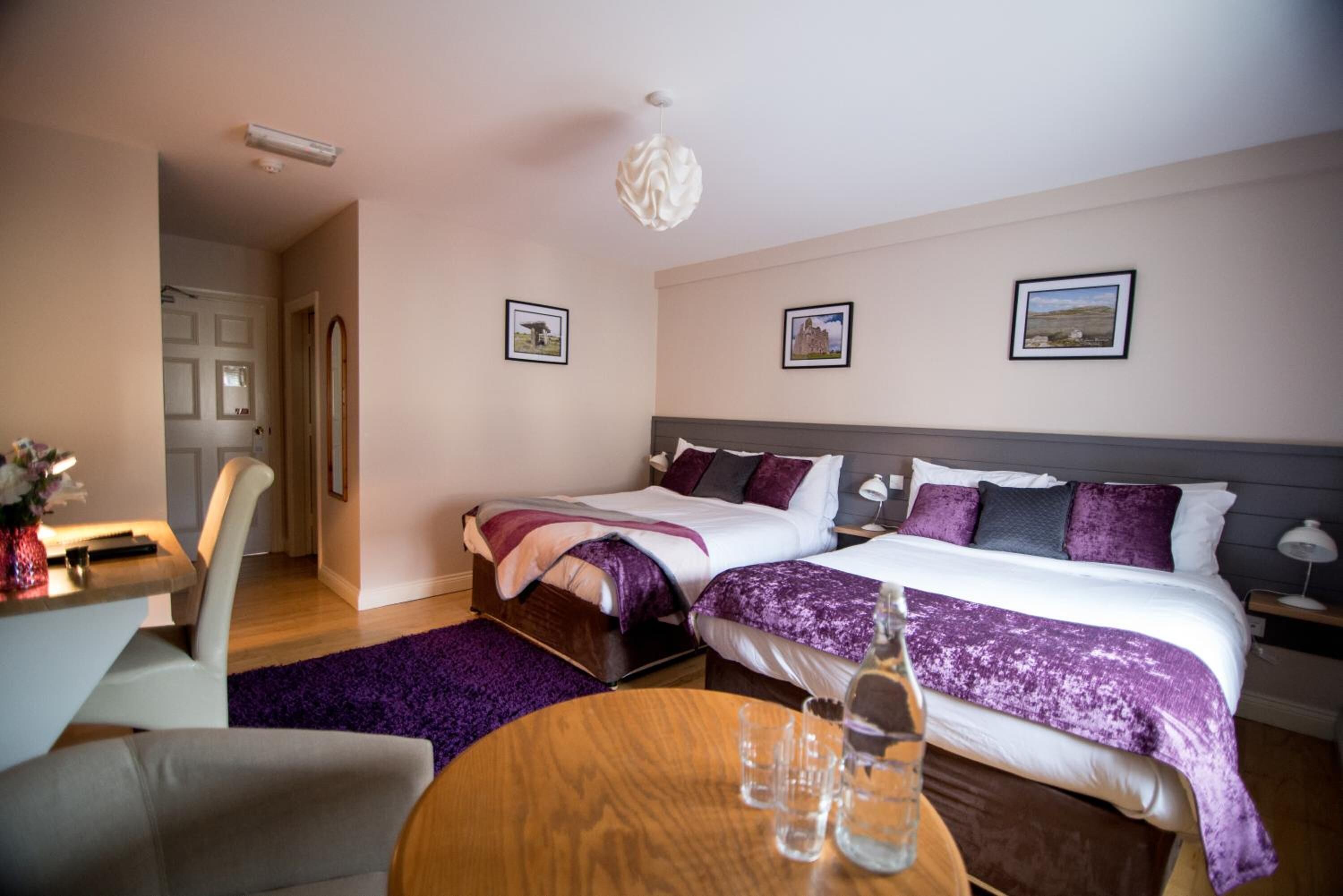 A hotel room with two beds, a wooden table, a purple rug, and framed pictures on the wall.