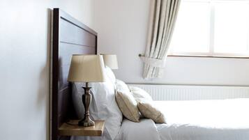 A neatly made bed with white linens and a wooden headboard, a bedside table with a lamp, and a window with curtains.