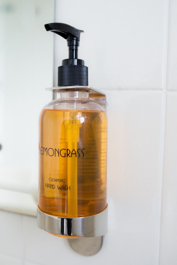 A hand wash dispenser with a pump, labeled "LEMONGRASS CLEANSING HAND WASH," mounted on a white tiled wall.