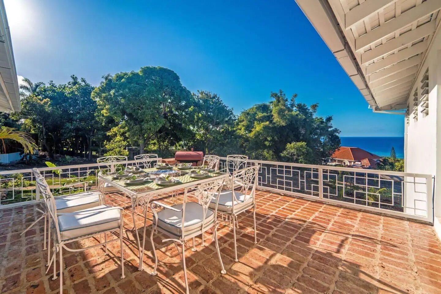 A balcony with a table set for dining, surrounded by white metal railings, overlooking a scenic view of trees and a body of water.