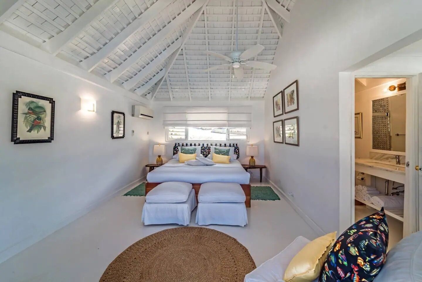 Un dormitorio con techo alto, dos camas, un ventanal grande y un baño con espejo y lavamanos.