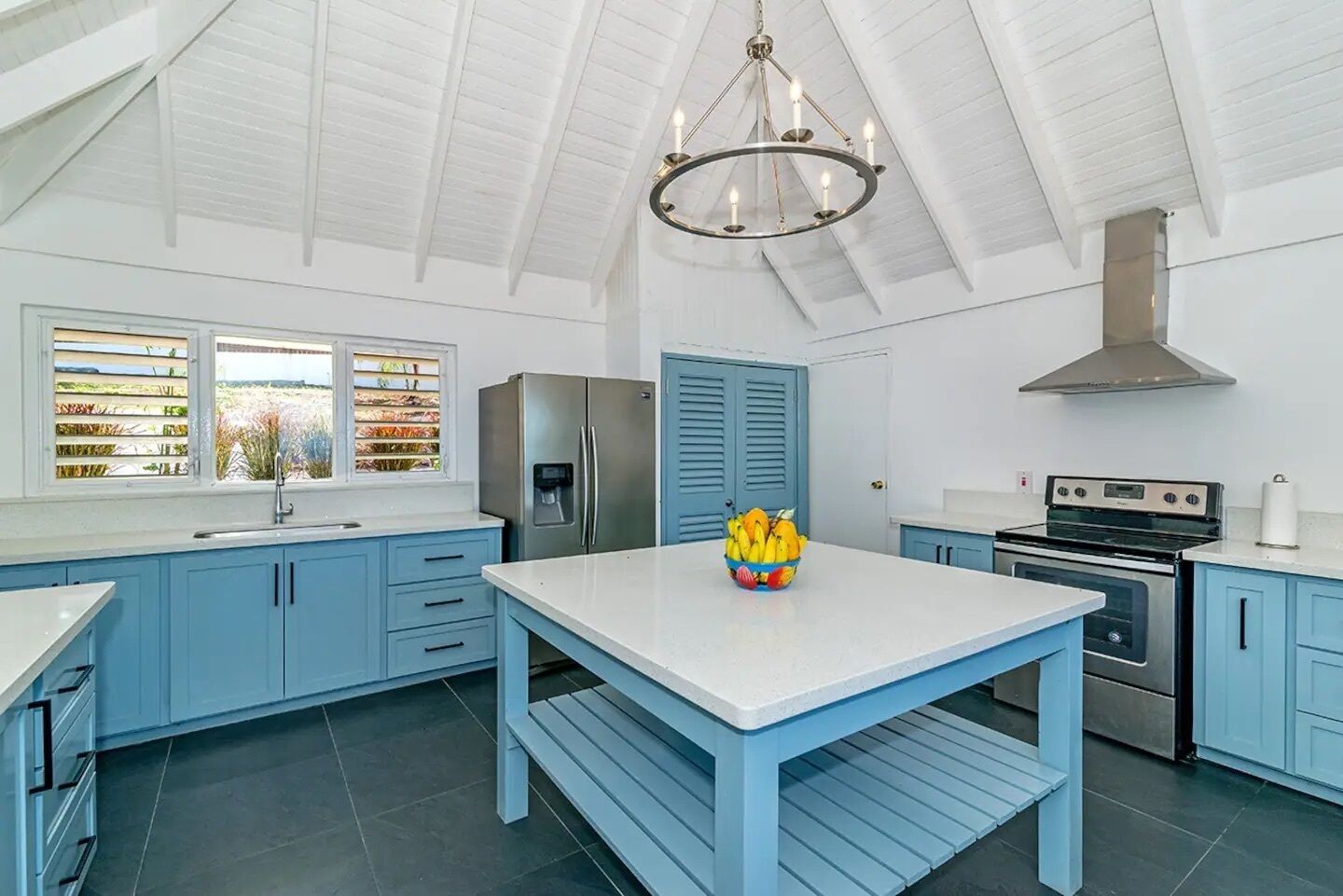 A modern kitchen with blue cabinets, a white countertop, a stainless steel refrigerator, a gas stove, and a chandelier.