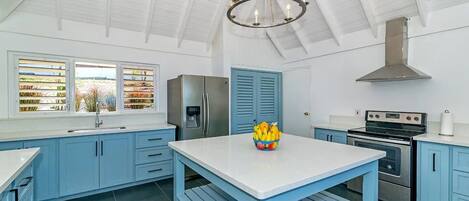 A modern kitchen with blue cabinets, a white countertop, a stainless steel refrigerator, a gas stove, and a chandelier.