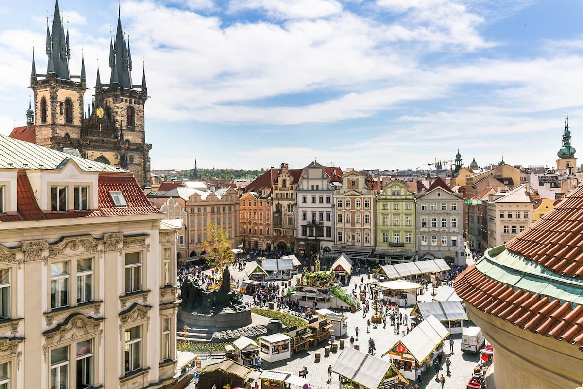 A historic city square with traditional buildings, a prominent church tower, and a central monument.