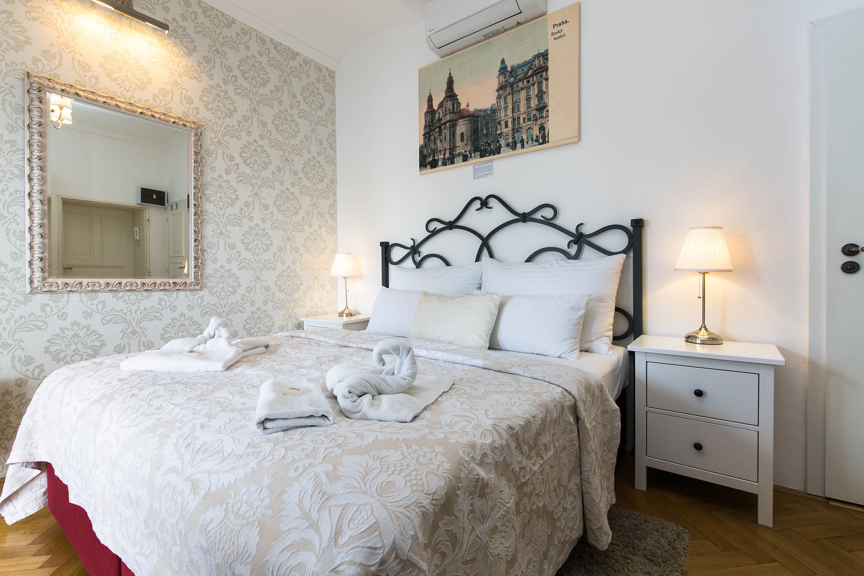 A neatly made bed with a patterned bedspread, a large mirror, a nightstand with a lamp, and a framed picture on the wall.