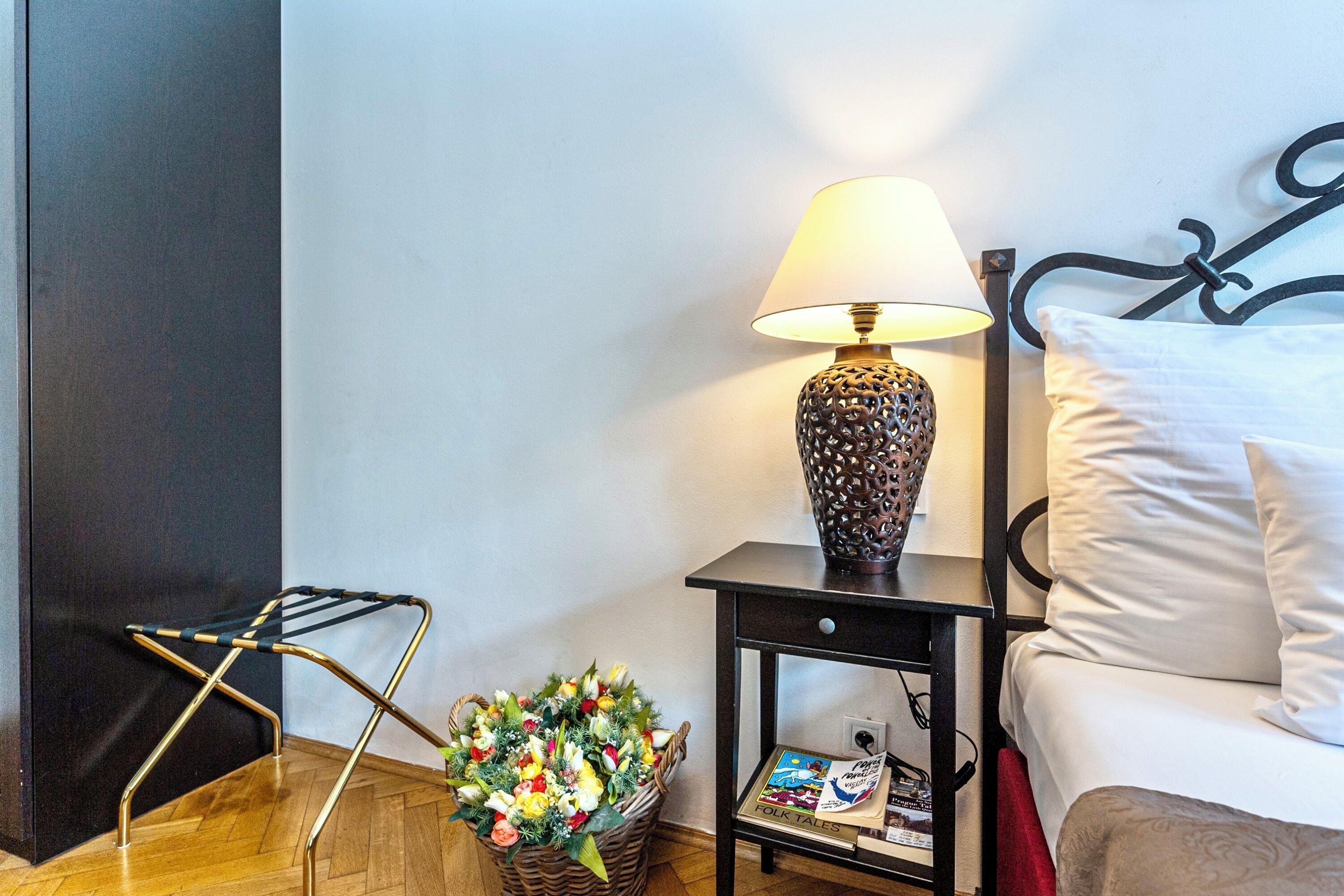 A neatly arranged hotel room with a bed, a bedside table with a lamp, a small shelf with books, and a basket of flowers.