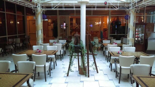 A spacious indoor dining area with multiple tables and chairs, a central plant, and a ceiling with a grid-like structure. - Hotel Kralj Tomislav (Nova Gradiska)