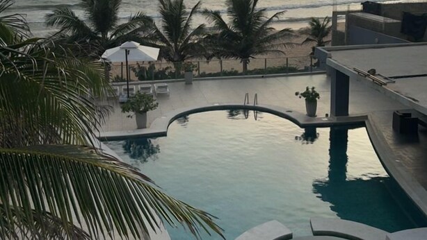 A pool area with a view of the ocean, palm trees, and a cloudy sky.