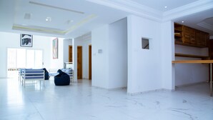 A spacious living area with a white marble floor, a white bench, and a blue bean bag chair. There are wooden cabinets and a small window on the right.