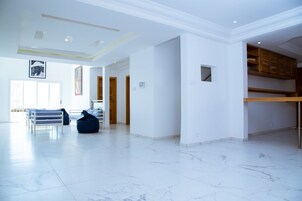 Amplia sala de estar con piso de mármol blanco, un banco blanco y un sillón de peluche azul. Se observan armarios de madera y una pequeña ventana a la derecha.