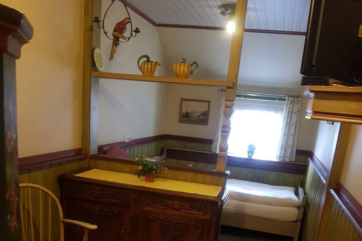 Ein kleiner Raum mit einer hölzernen Bank, einem Tisch mit gelber Tischdecke, einem Stuhl und einem Bett.