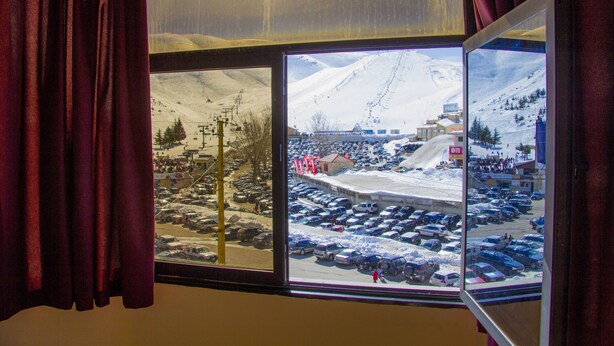 A view through a window with curtains, featuring a snowy parking lot and a mountainous landscape with buildings and ski lifts.