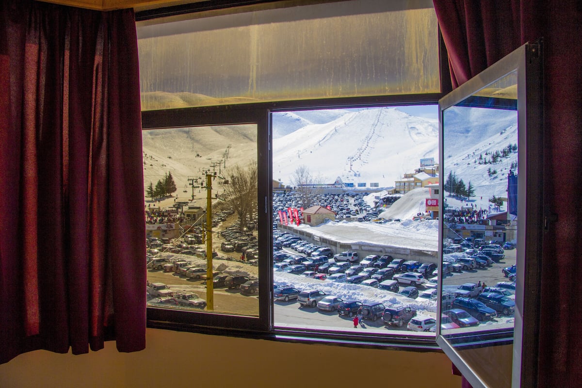 Vista a través de una ventana con cortinas, mostrando un estacionamiento nevado y un paisaje montañoso con edificios y remontes.